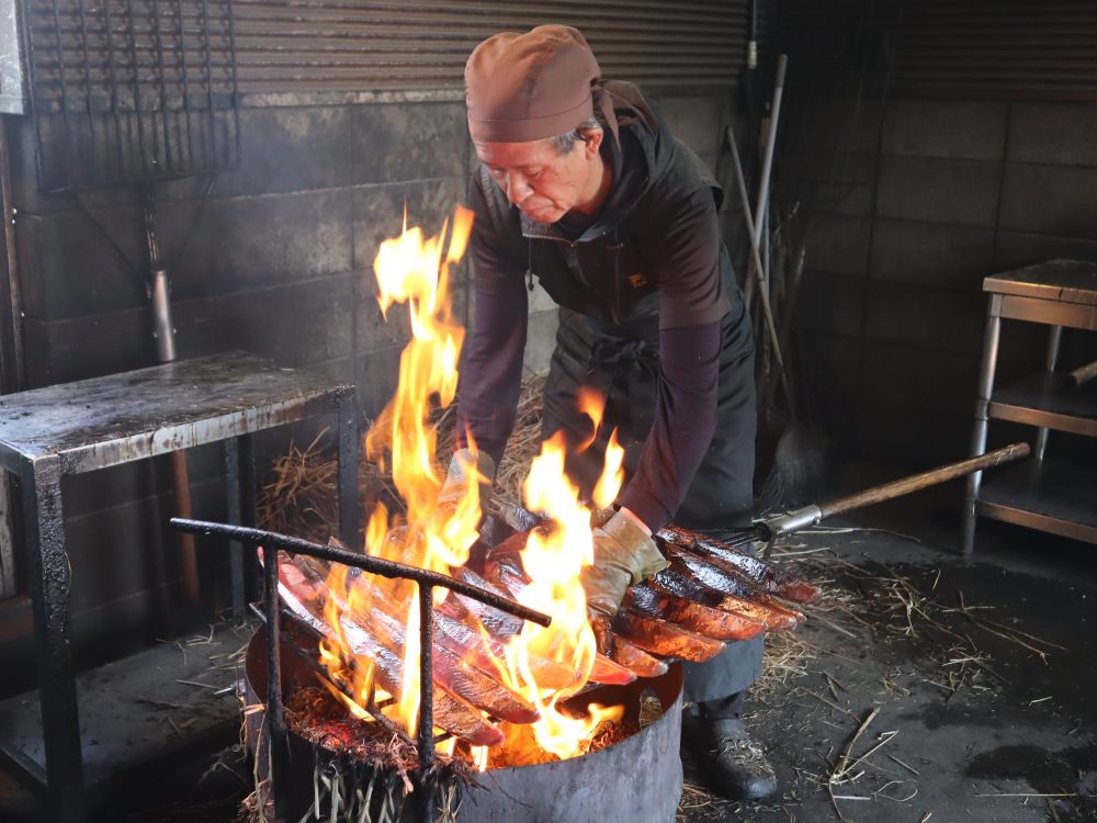 ドラム缶の上に網をのせ鰹のタタキを焼いている様子。炎が上がる中に手を入れ、職人が鰹を動かしている。