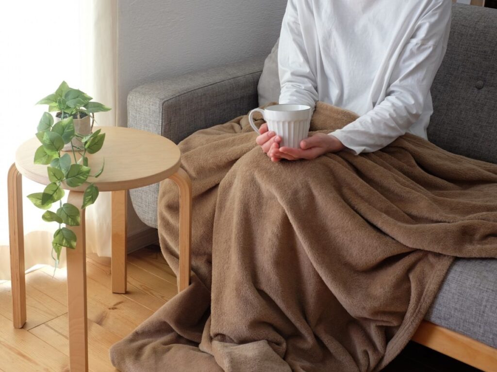 人がソファで毛布を掛けて座り、飲み物を飲んでいる