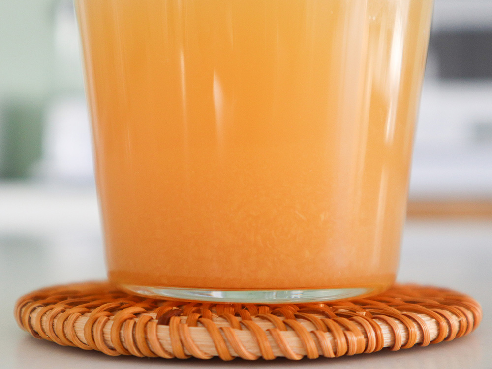 ゴールドパックの商品である『食べるアップルジュース』をコップに注ぎ、横から撮った写真。オレンジがかった黄色いりんごのジュースの中に、沈殿している果肉を確認できる。