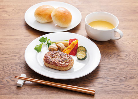 白い皿にのった黒豚と鶏のハンバーグの写真 奥にパンとスープがおいてある