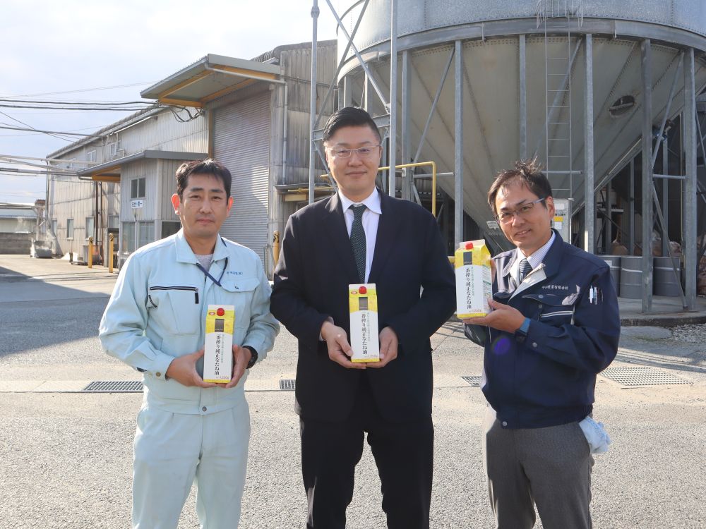 『自然派Style一番搾り純正なたね油』を持って工場の前に立つ平田産業の社長、柳田さん、安達さん