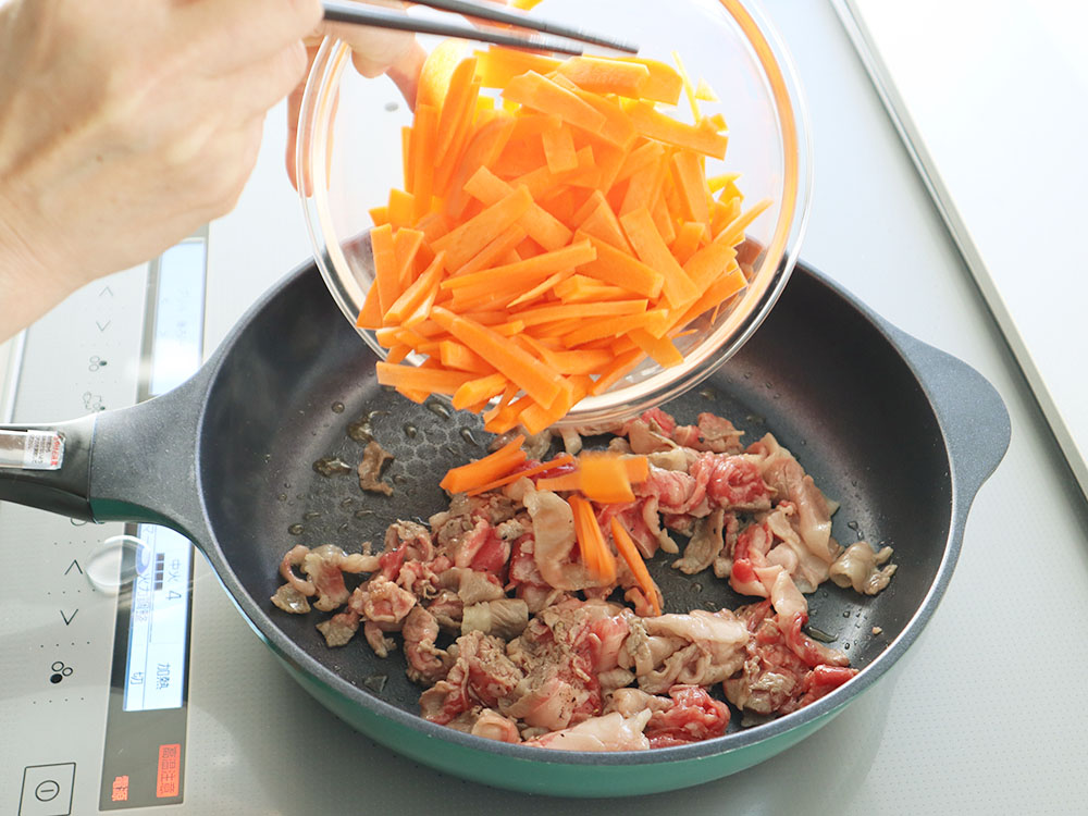 肉が炒められているフライパンへ細切りにしたにんじんを加えるシーン