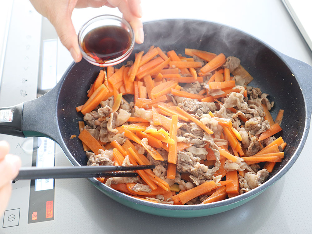 牛肉とにんじんが炒められているフライパンへめんつゆを回しかけるシーン